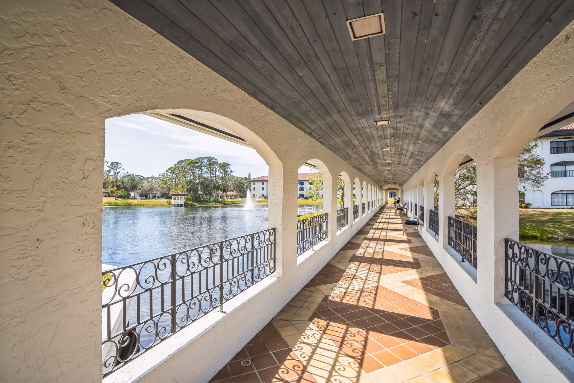 Covered outdoor walkway with arched openings and decorative wrought iron railings overlooking a lake with a fountain and gazebo, with buildings and trees in the background.