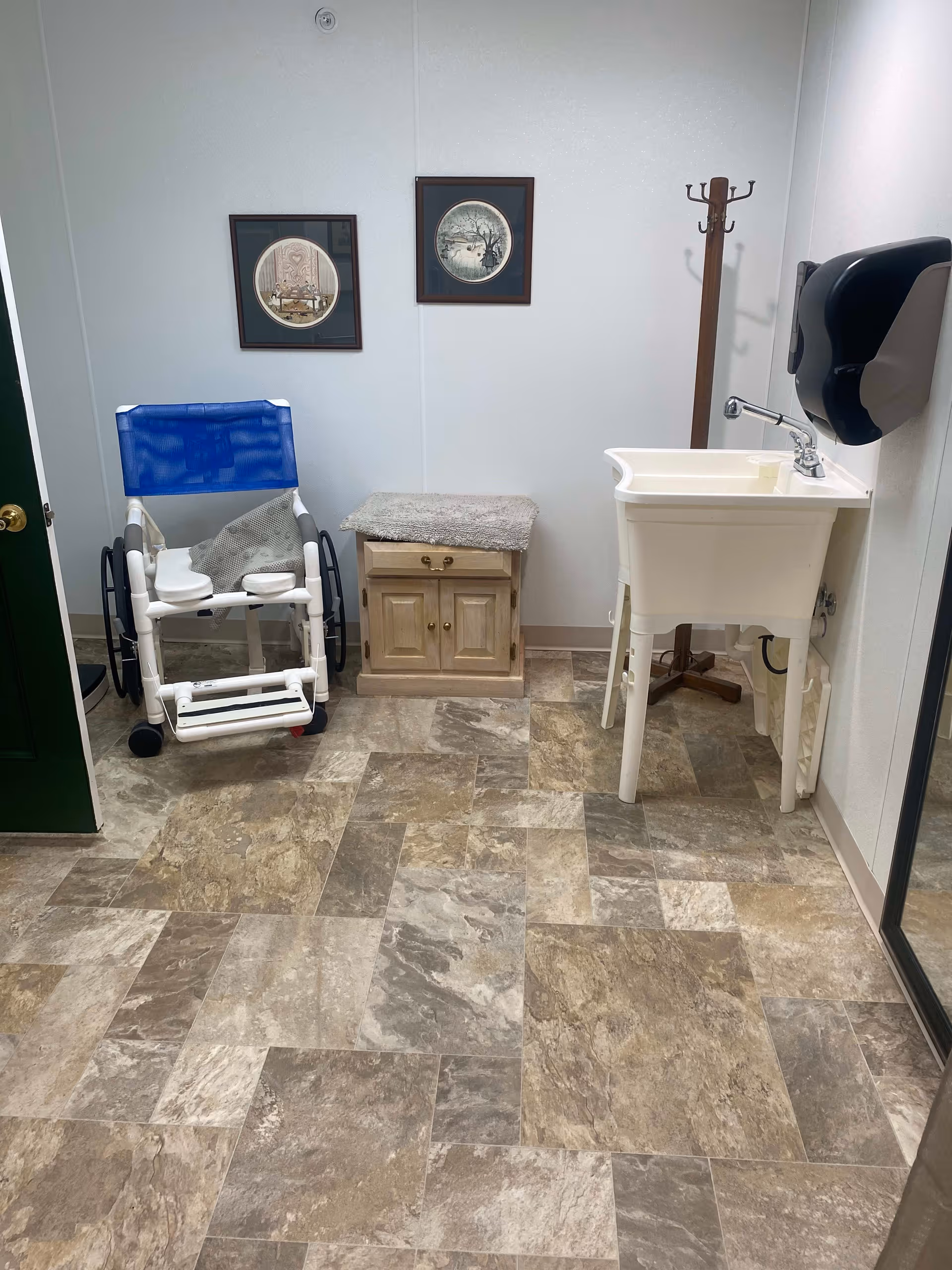 A small room with a blue and white wheelchair, a wooden cabinet with a cushioned top, a white utility sink with a faucet, a wall-mounted black paper towel dispenser, a wooden coat rack, and two framed pictures hanging on a white wall.