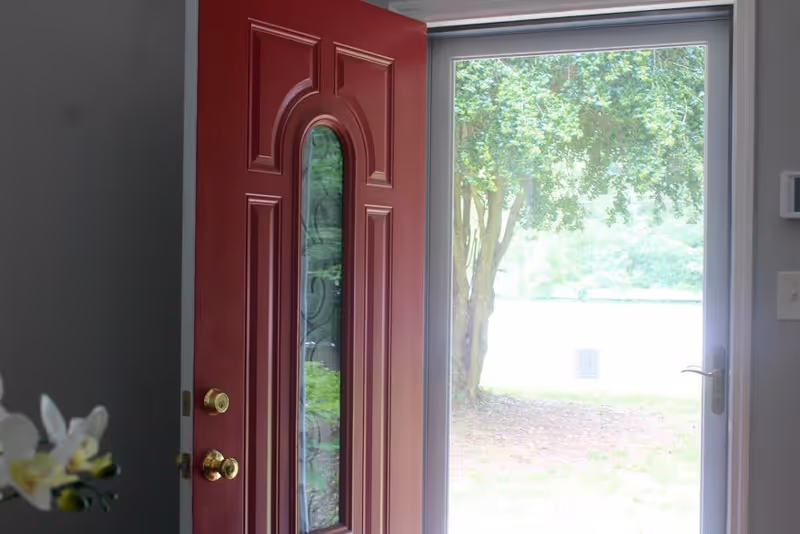 An open red front door with decorative glass panels, leading to an outdoor area with green trees and grass visible through a large glass storm door.