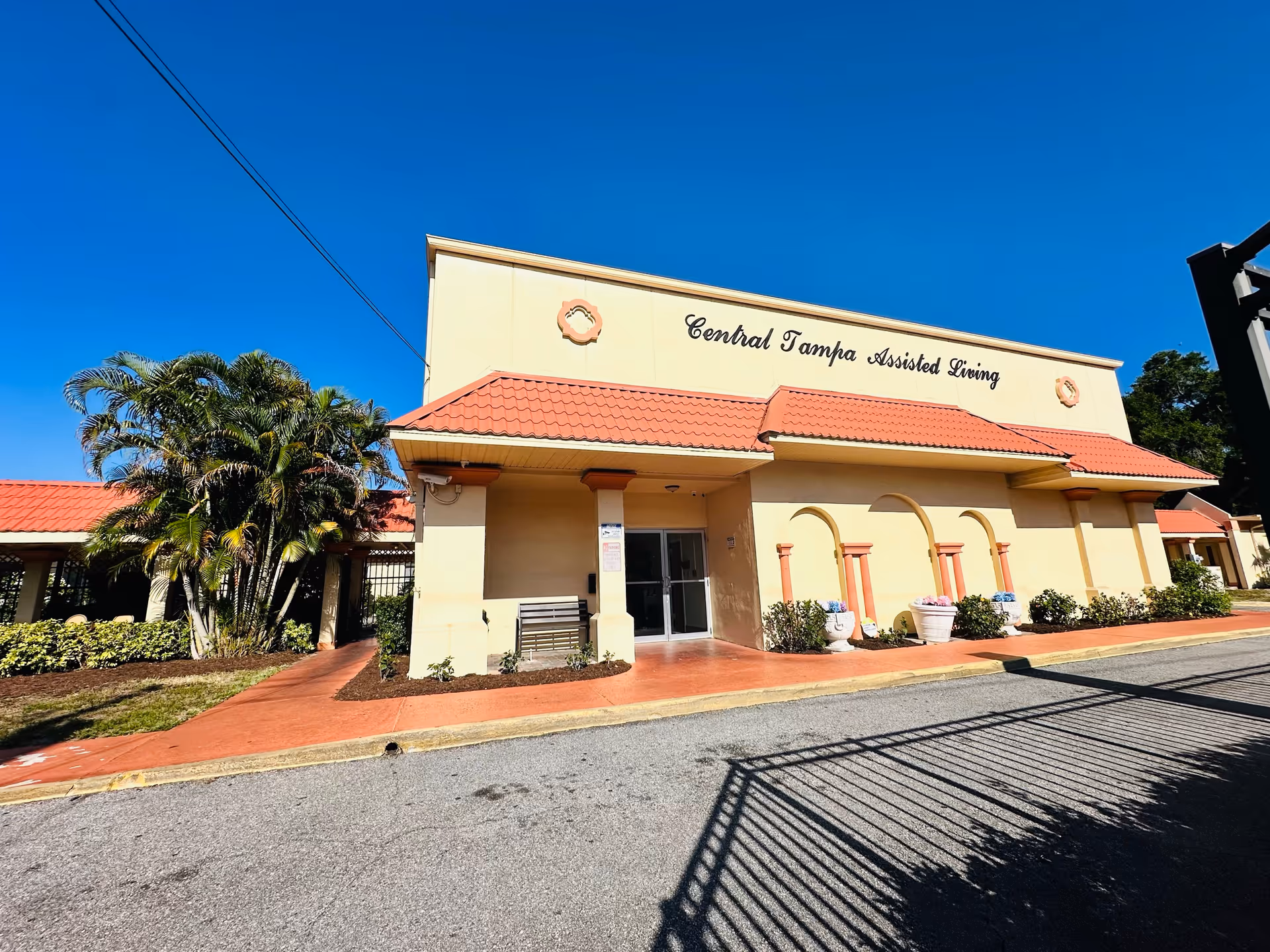 Exterior view of a building with a beige facade and red tiled roof under a clear blue sky. The building has a sign that reads 'Central Tampa Assisted Living'. There are palm trees and shrubs along the walkway leading to the entrance with glass doors.