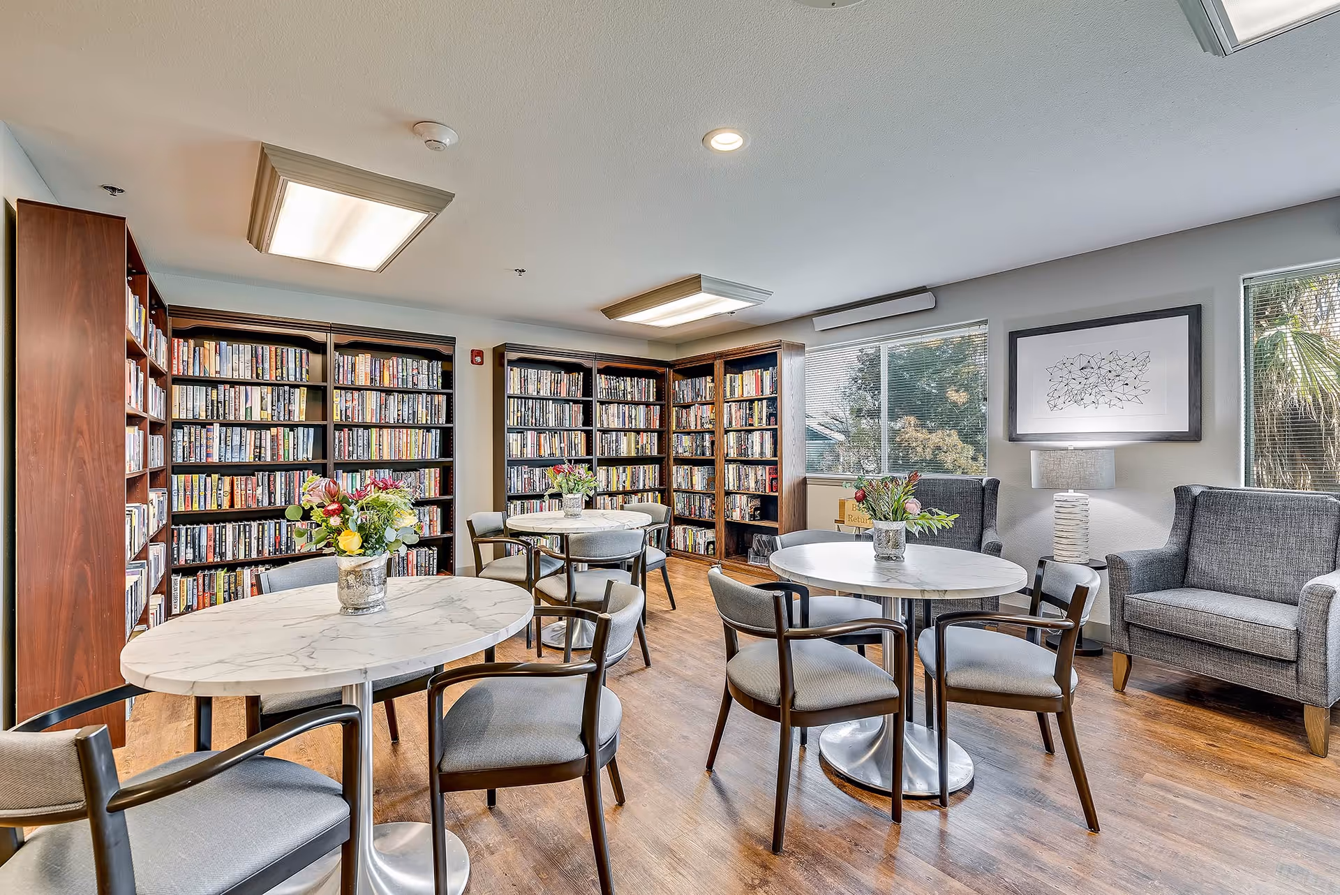 Bright communal library room with round marble tables, chairs, tall bookshelves and lounge chairs by windows.