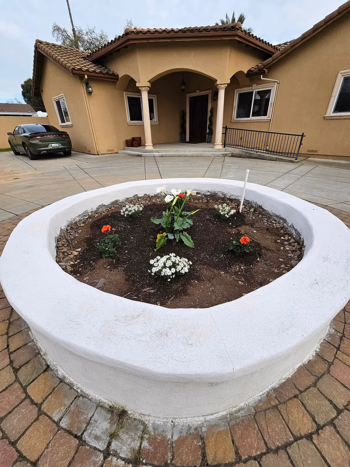 A circular raised flower bed with various small flowers planted inside, situated in front of a beige stucco building with a tiled roof and an arched entrance supported by columns. A green car is parked to the left on a concrete driveway.