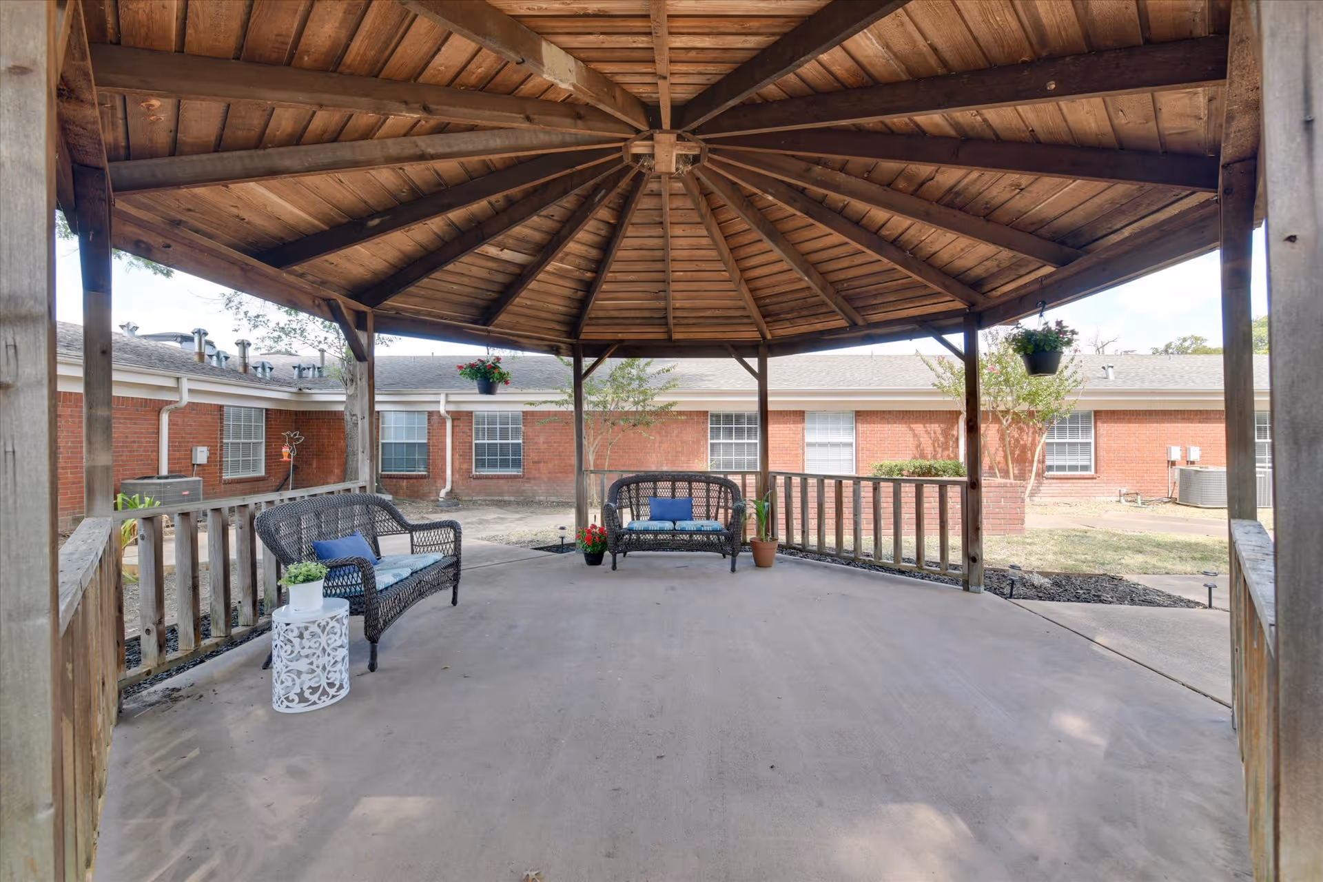 A wooden gazebo with a pitched roof in an outdoor courtyard area. Inside the gazebo are two black wicker benches with blue cushions, a small white decorative side table with a potted plant, and two hanging flower pots. The background shows a brick building with several windows and some landscaping.