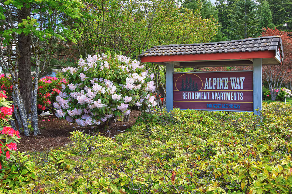 Entrance sign reading "Alpine Way Retirement Apartments" surrounded by flowering shrubs and trees.