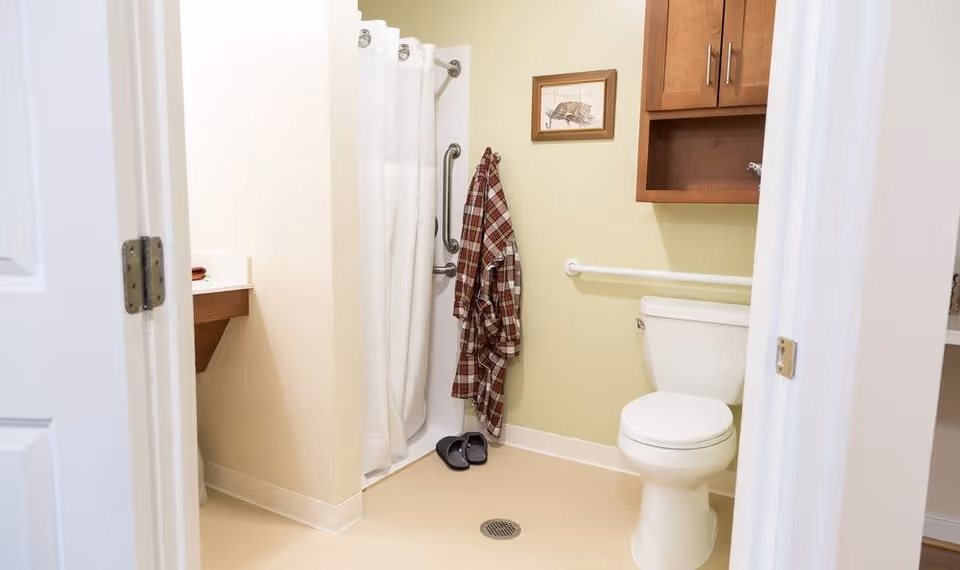 A clean and accessible bathroom with a white toilet, a shower with a white curtain, a plaid robe hanging on a hook, a pair of slippers on the floor, a wooden cabinet mounted on the wall, and a framed picture above the toilet.