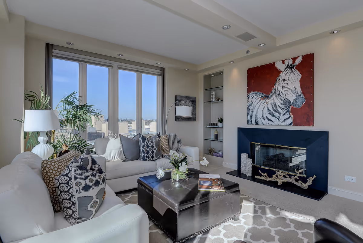 Bright modern living room with white sofas and patterned pillows, a central black ottoman, large windows, and a fireplace topped by a zebra painting.