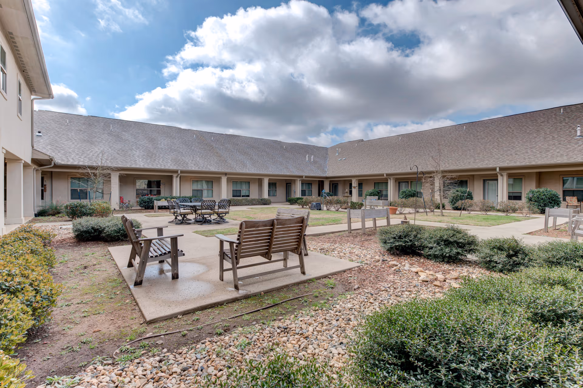 Outdoor courtyard area of a senior living facility with wooden benches, chairs, tables, and landscaped bushes and plants under a partly cloudy sky.