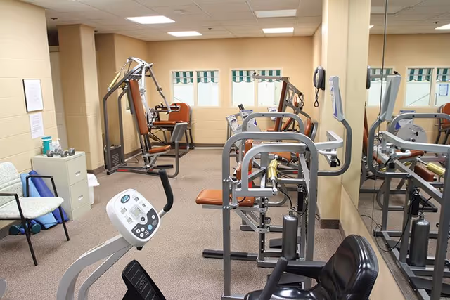 Exercise room with several strength-training machines, a recumbent bike, mirrored wall and seating.