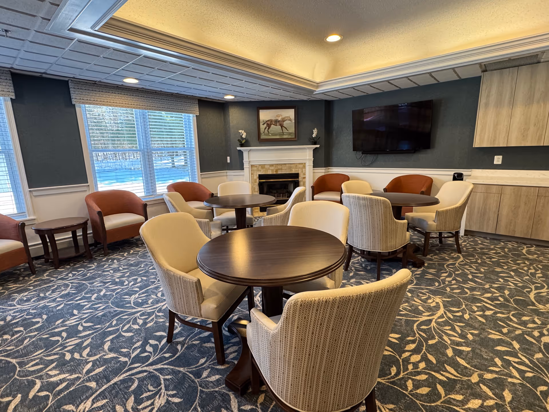 A cozy common area with multiple round tables surrounded by cushioned chairs. The room features a patterned carpet, large windows with blinds, a fireplace with a horse painting above it, a wall-mounted flat screen TV, and built-in cabinets along one wall.