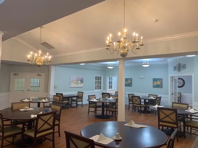 A dining room with several round tables and chairs arranged neatly. Each table has white folded napkins and small cups placed on it. The room has light blue walls, wooden flooring, and two ornate chandeliers hanging from the ceiling. There are paintings on the walls and double doors with glass panels in the background.
