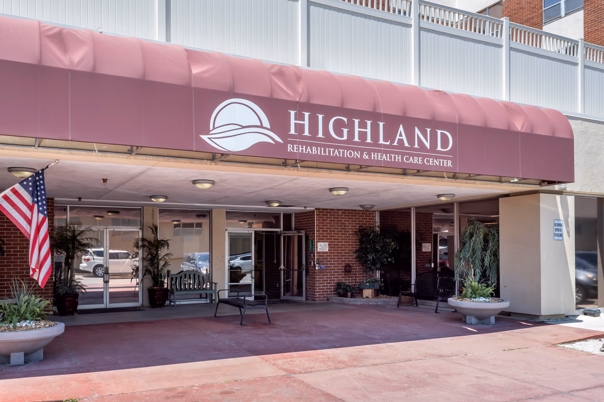 Entrance of Highland Rehabilitation & Health Care Center with a maroon awning, benches, planters, and an American flag.