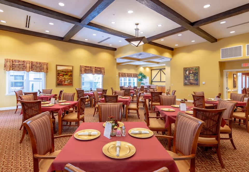 A spacious dining room with multiple tables covered in red tablecloths, each set with gold plates and white napkins. The room has yellow walls, patterned carpet, wooden chairs with striped upholstery, and windows with valances. Ceiling beams and recessed lighting add to the warm ambiance.