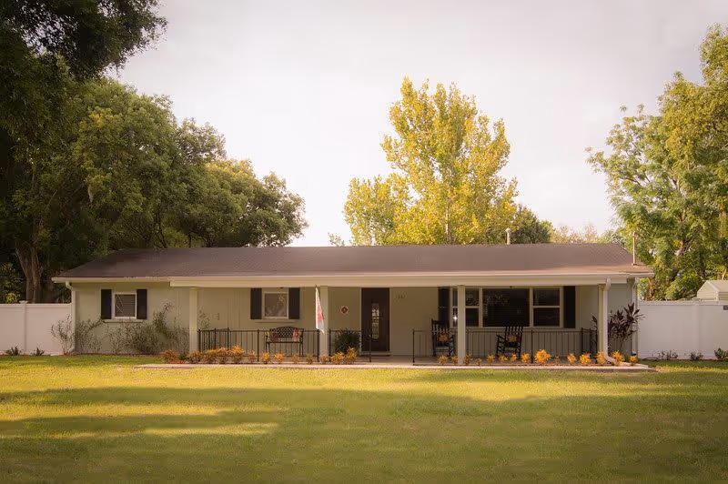 Single-story ranch-style building with a covered front porch, rocking chairs, and a large lawn with trees behind.