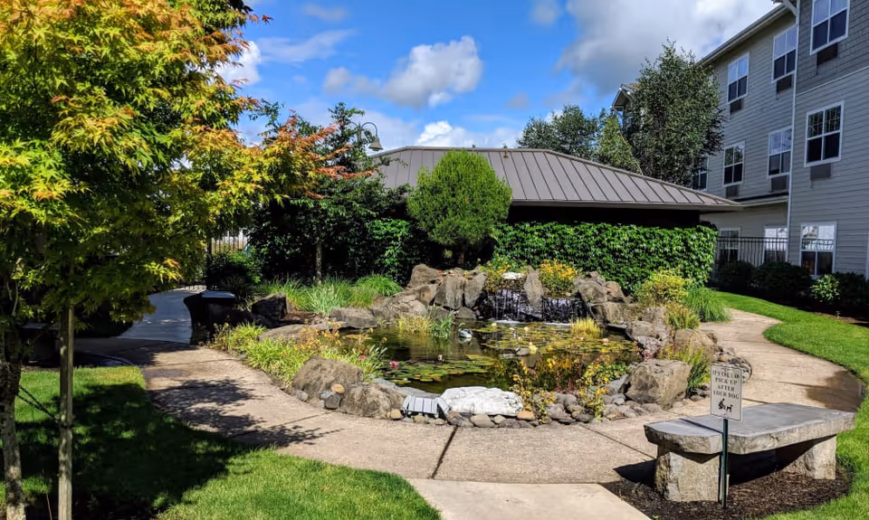 Courtyard with a rock-lined pond and small waterfall, paved paths, bench, and adjacent senior living building under a blue sky.