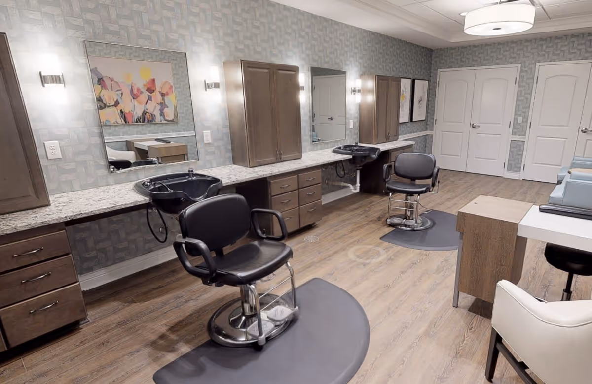 Interior view of a salon area in a senior living facility with two black salon chairs positioned in front of sinks and mirrors. The room features wood flooring, light gray patterned wallpaper, wooden cabinets, and modern lighting fixtures. There is a small desk with a white chair in the foreground and two closed white doors in the background.