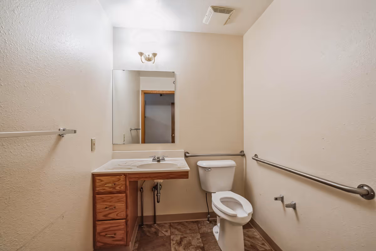 A small bathroom with beige walls and tiled floor featuring a white toilet with grab bars on both sides and a wooden vanity with a white sink and a mirror above it. There is a light fixture above the mirror and a towel bar on the left wall.