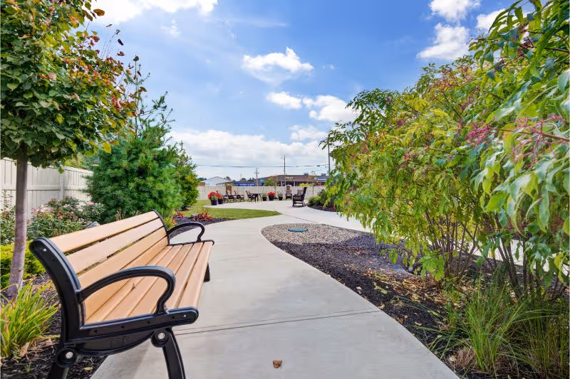 A curved concrete pathway in an outdoor garden area with a wooden bench with black metal armrests on the left side. The path is bordered by green shrubs and small trees, with a bright blue sky and some clouds overhead. In the distance, there are more benches and some buildings visible.