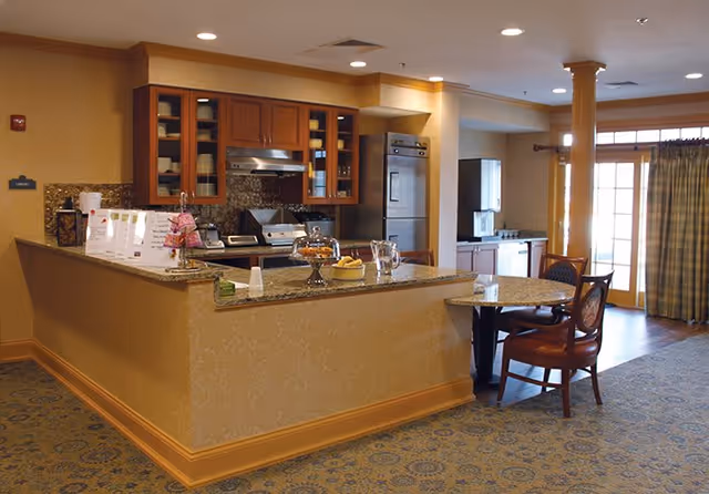 A warm and inviting kitchen area with wooden cabinets, a granite countertop island with baked goods and a pitcher on it, stainless steel appliances including a stove and refrigerator, and a small round table with two chairs. The room has recessed lighting, patterned carpet, and large windows with curtains allowing natural light to enter.