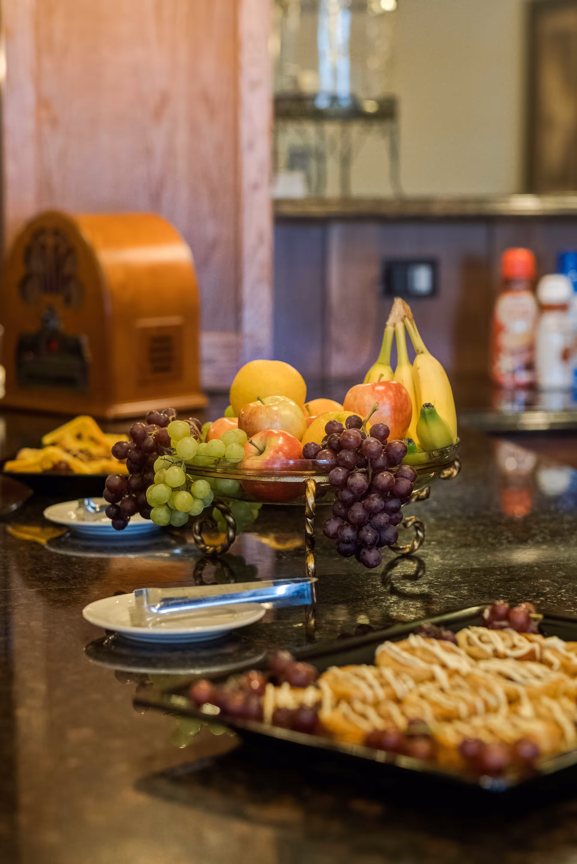 A kitchen counter with a decorative fruit bowl containing apples, bananas, grapes, and an orange. In the foreground, there is a plate with pastries topped with icing and grapes. In the background, there are condiment bottles and a vintage-style radio on the counter.
