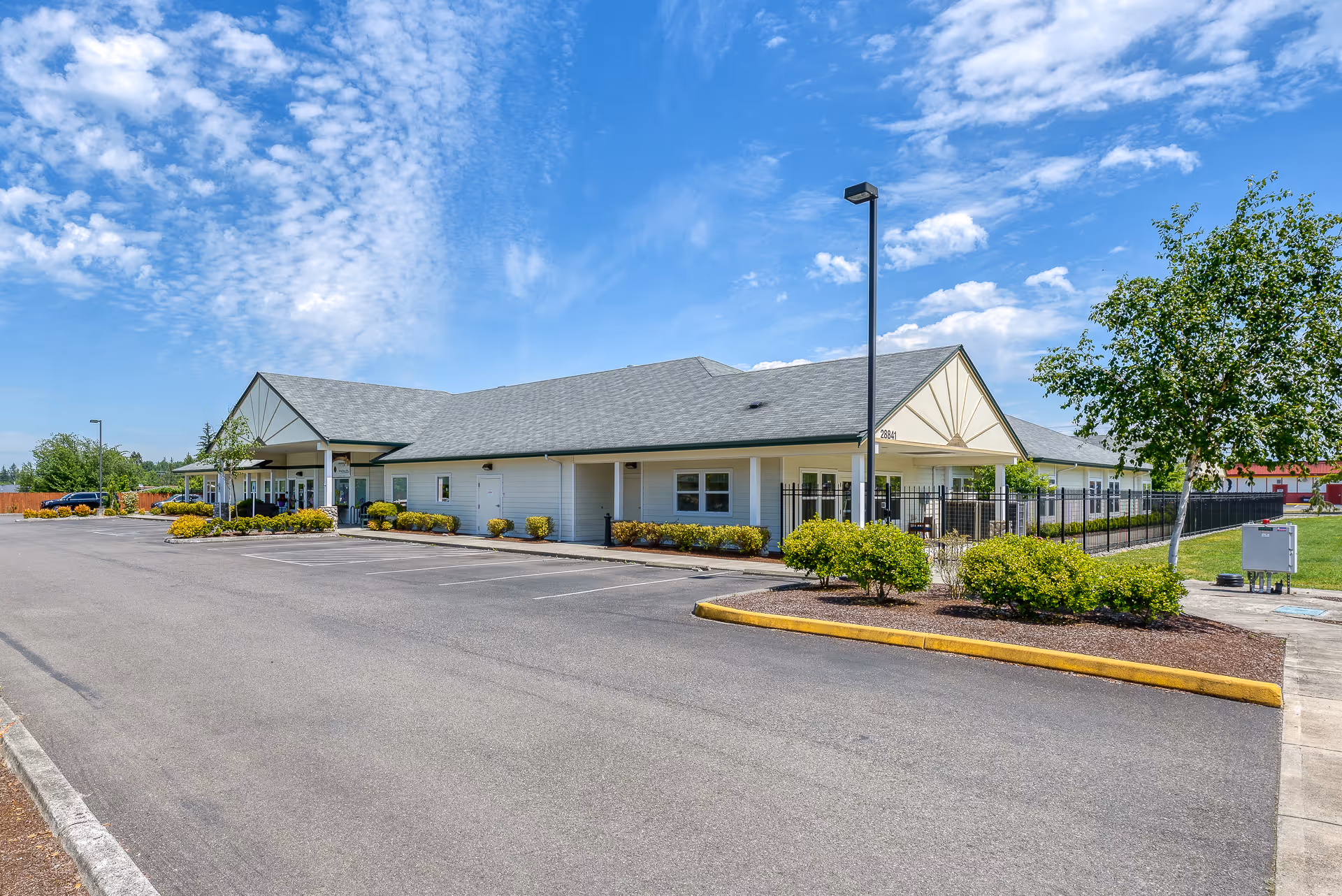 Exterior view of a single-story assisted living facility building with a gray roof, white siding, and a covered entrance. The building is surrounded by a parking lot, landscaped bushes, and a tree under a partly cloudy blue sky.