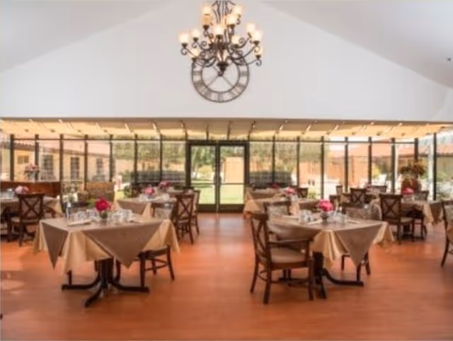 A bright dining room with multiple tables covered in beige tablecloths, each set with plates, glasses, and pink flower centerpieces. The room has large windows letting in natural light and a chandelier hanging from the ceiling. Outside greenery is visible through the windows.