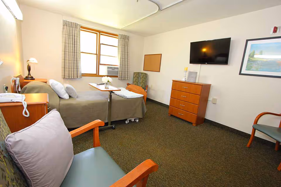 A senior living facility bedroom with a single bed covered in a gray blanket, a bedside table with a lamp and telephone, a small overbed table with a flower vase, two armchairs with cushions, a wooden dresser with a flat-screen TV mounted above it, a window with curtains, and a framed picture on the wall.