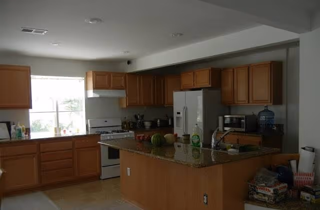 Open kitchen with wooden cabinets, a granite-topped island with sink, refrigerator, stove, and a window letting in natural light.