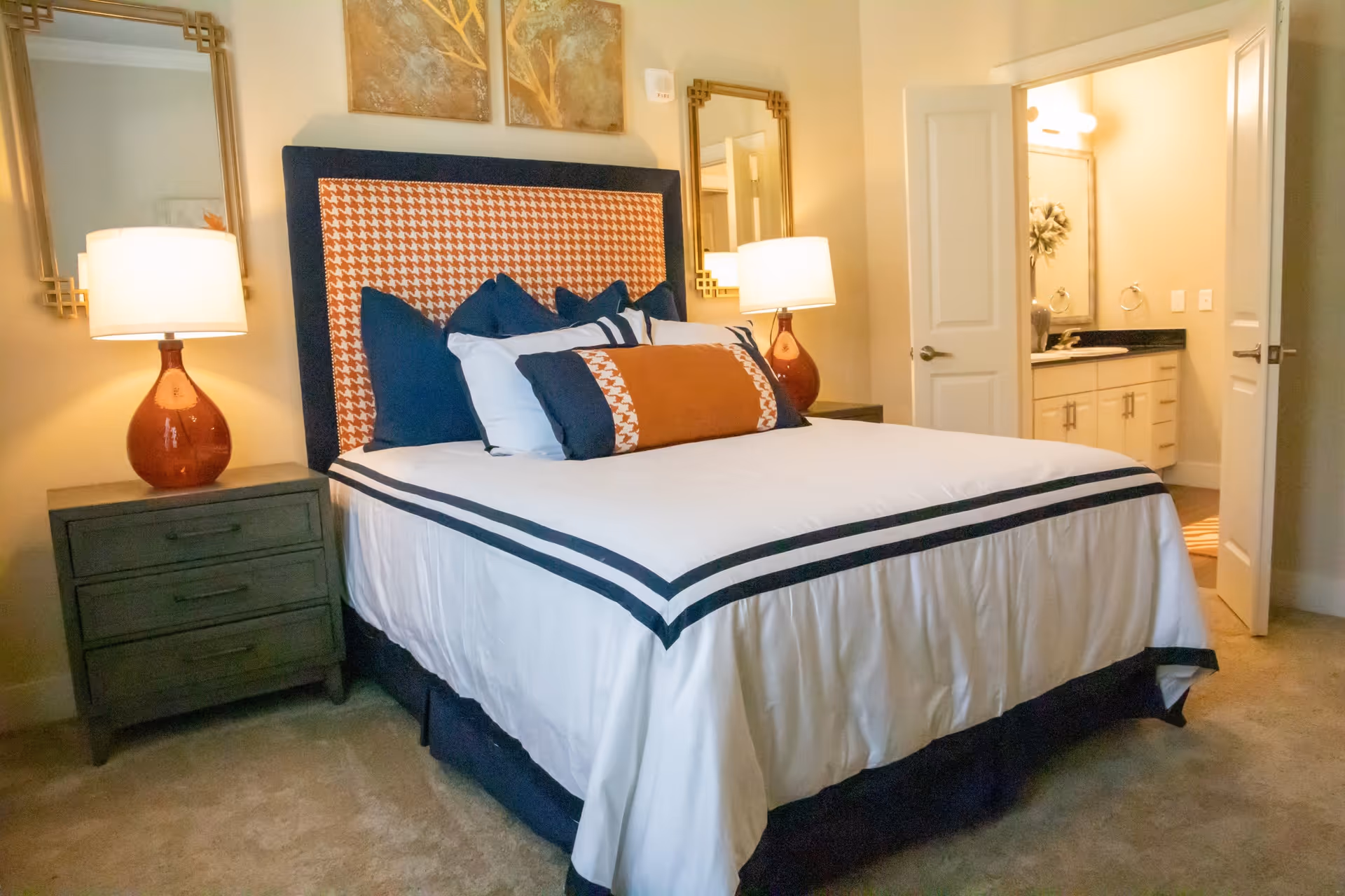 A neatly made bed with a patterned headboard and multiple pillows in a bedroom. There are two bedside tables with matching lamps on either side of the bed. Two decorative mirrors hang on the wall above the bedside tables. An open door reveals a bathroom with a sink, countertop, and a large mirror.