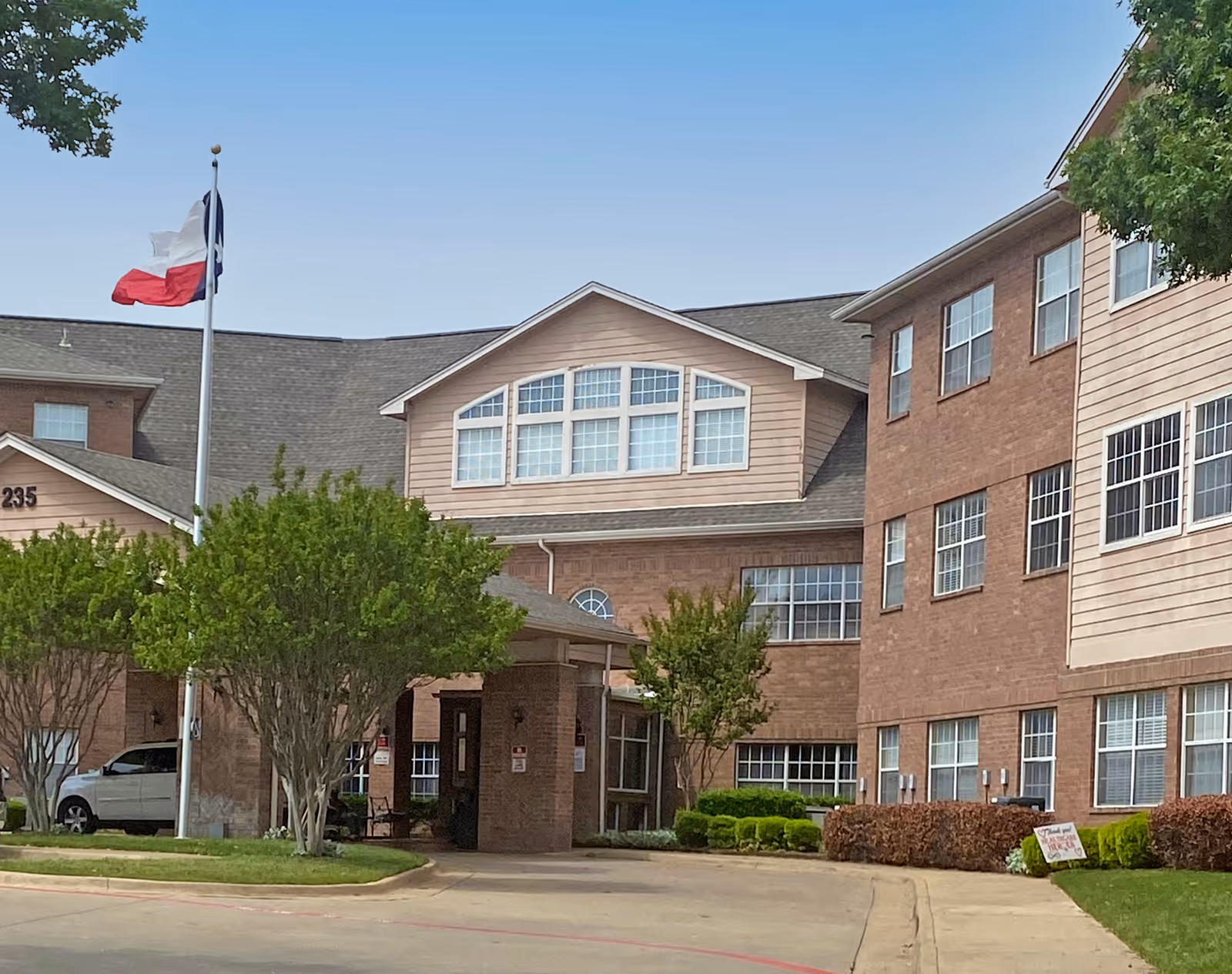 Exterior view of Morada Cedar Hill facility showing a multi-story brick building with numerous windows, a covered entrance, a flagpole with the Texas state flag, and landscaped greenery including trees and bushes under a clear blue sky.