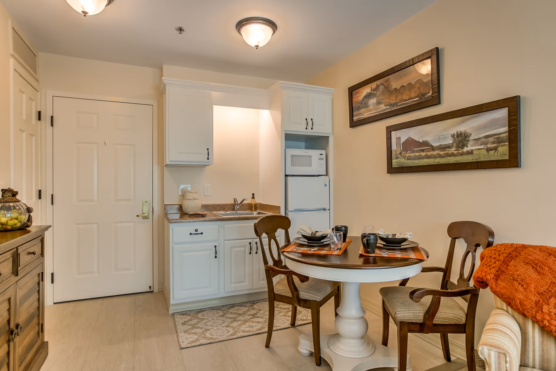 A small dining and kitchen area in a retirement community apartment. The kitchen features white cabinets, a sink, a microwave, and a refrigerator. A round dining table is set for two with plates, bowls, glasses, and mugs. Two wooden chairs with cushions are placed around the table. The wall has two framed landscape paintings, and a cozy orange blanket is draped over a nearby sofa.