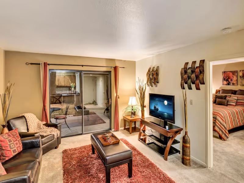 A cozy living room with a leather sofa and armchair, a red area rug, a wooden coffee table, and a TV on a wooden stand. There are decorative wall hangings and a lamp on a side table. Sliding glass doors lead to an enclosed patio area. A doorway on the right shows a bedroom with a striped bedspread and floral artwork on the wall.