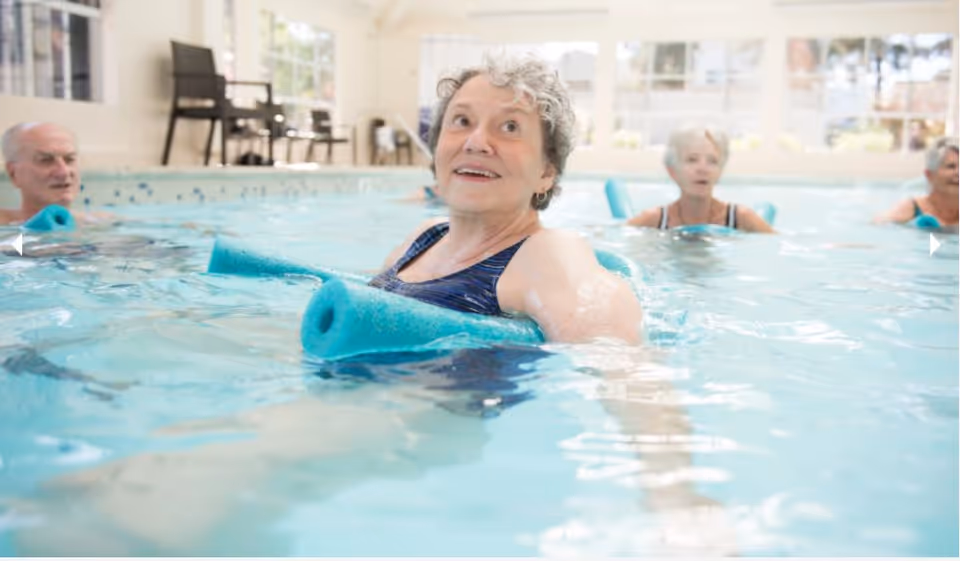 A group of elderly people participating in a water exercise class in an indoor swimming pool, using blue pool noodles for support. The room is bright with large windows letting in natural light.