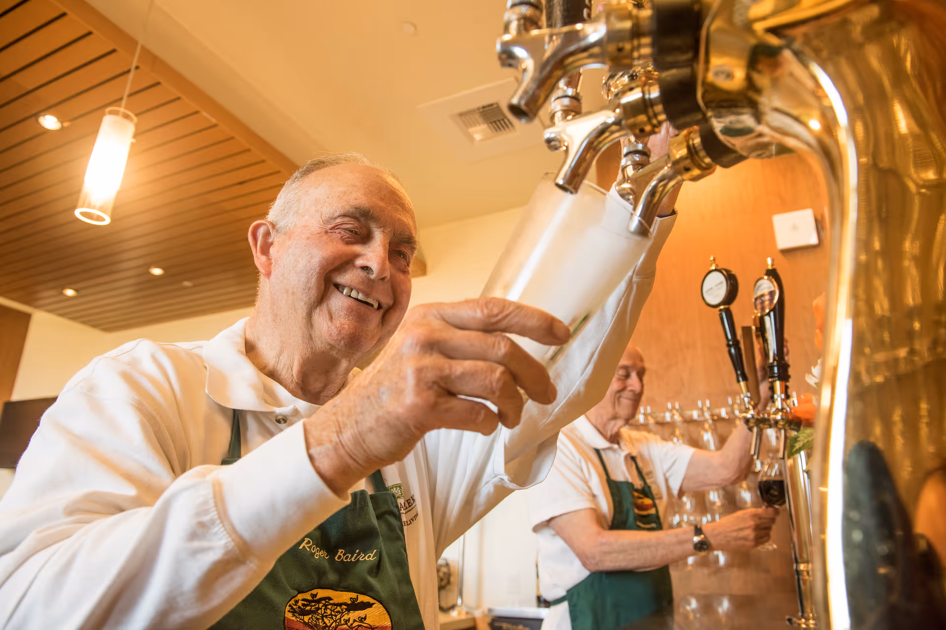 Two elderly men wearing aprons are smiling and pouring drinks from taps in a well-lit indoor setting with wooden ceiling panels.