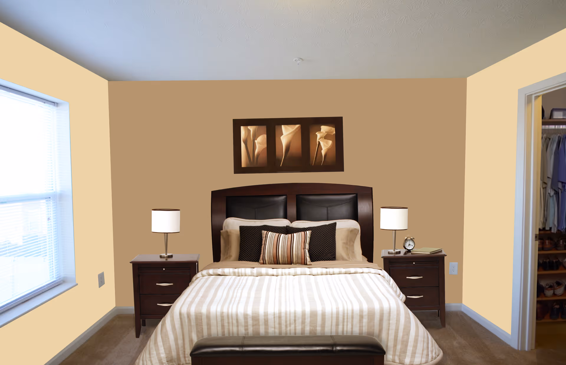 A neatly made bed with striped beige and white bedding, two dark wood nightstands with lamps, and a framed triptych of floral artwork above the headboard in a bedroom with beige walls and a window on the left side. A closet with hanging clothes and shoes is visible through an open door on the right.