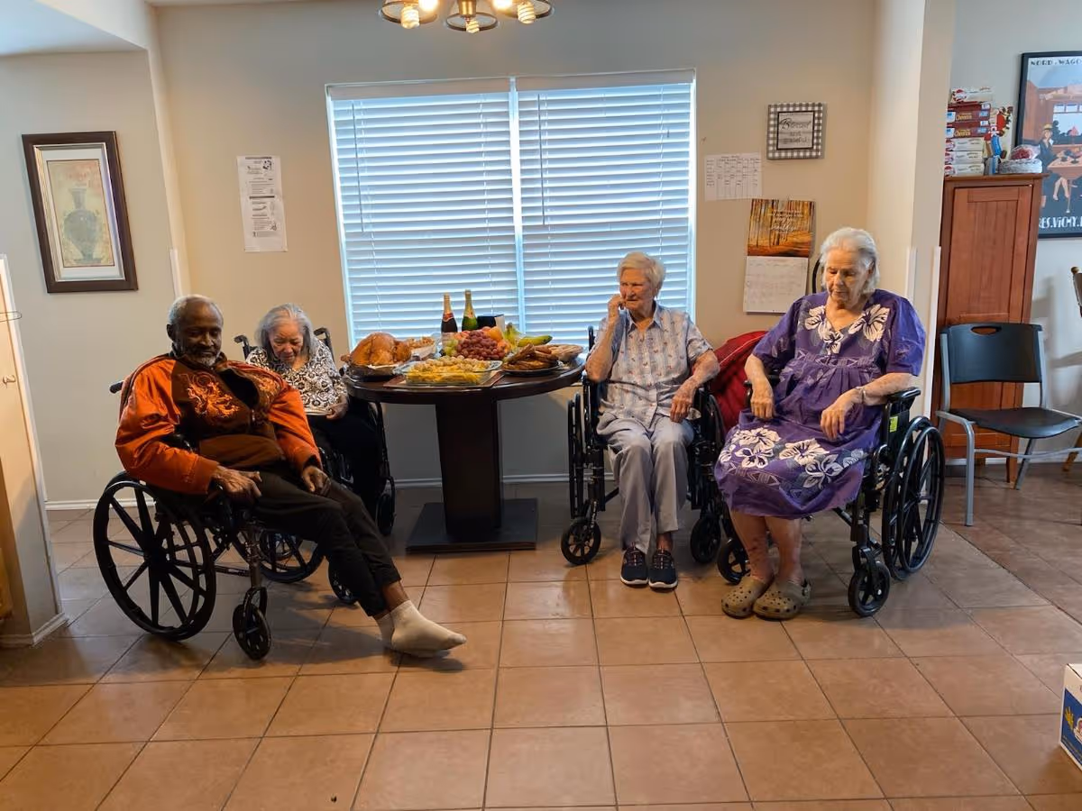 Four elderly residents in wheelchairs seated in a communal room with a table of food and drinks in front of a window.