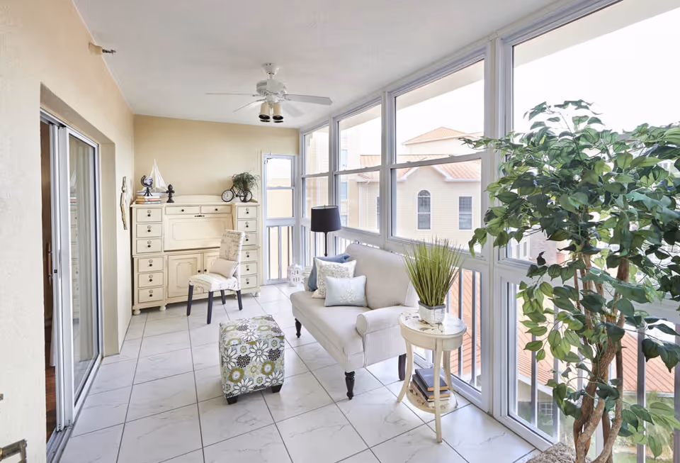 A bright enclosed balcony or sunroom with large windows showing a view of neighboring buildings. The room features a white cushioned loveseat with decorative pillows, a floral patterned ottoman, a small round side table with a potted plant, a cream-colored cabinet with drawers, a chair, a floor lamp, and a large leafy indoor plant. The floor is tiled in white, and a ceiling fan is mounted above.
