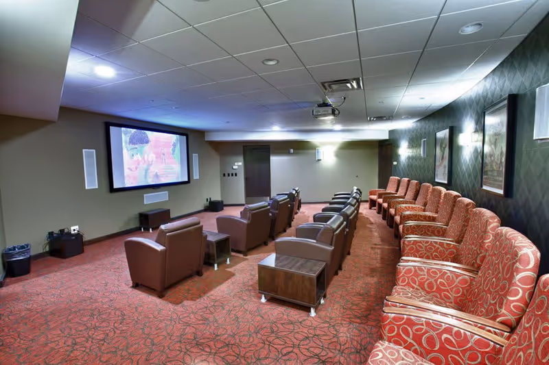 Small theater-style screening room with rows of recliner chairs and patterned seats facing a large wall-mounted projection screen.
