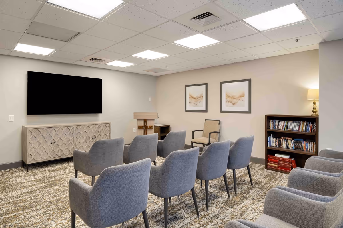 A small meeting or media room with two rows of gray upholstered chairs facing a large flat-screen TV mounted on a light gray wall. Below the TV is a decorative cabinet. The room has a patterned carpet, two framed abstract artworks on the wall, a wooden podium, a single armchair, a bookshelf filled with books and DVDs, and a lamp on a side table.