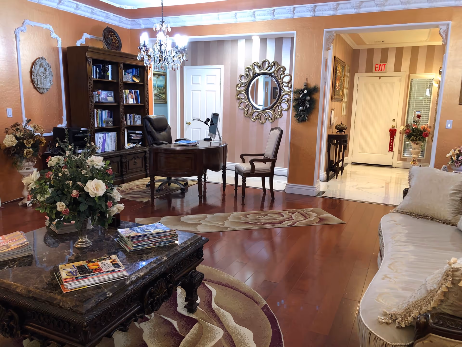 A warmly decorated living room area with polished wooden floors, a beige sofa with decorative pillows, a dark ornate coffee table with magazines and a floral arrangement, a wooden desk with a computer and chair, a bookshelf filled with books, a chandelier hanging from the ceiling, and a hallway leading to a door with an exit sign above it.