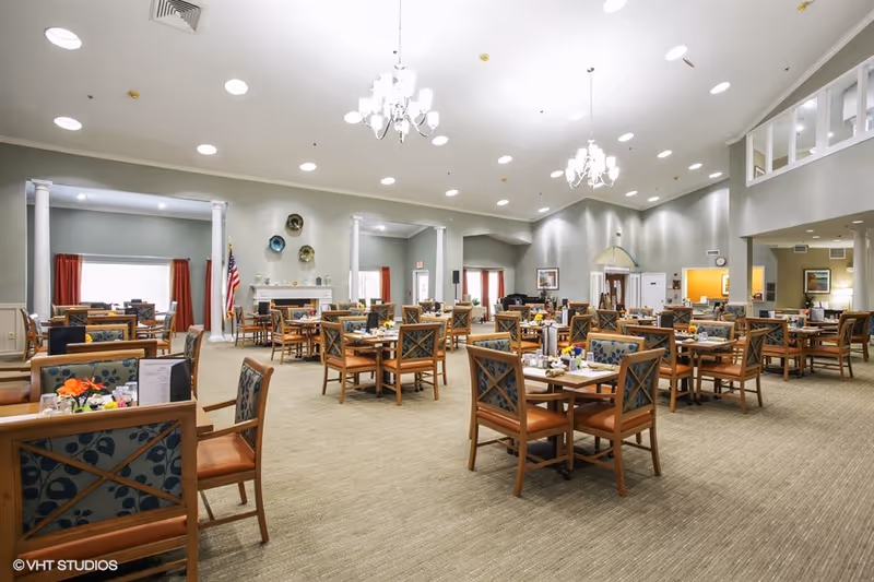 A spacious dining room with multiple wooden tables and chairs arranged neatly. The room features high ceilings with chandeliers, large windows with red curtains, and decorative wall plates above a fireplace. The setting is bright and welcoming, suitable for communal dining.