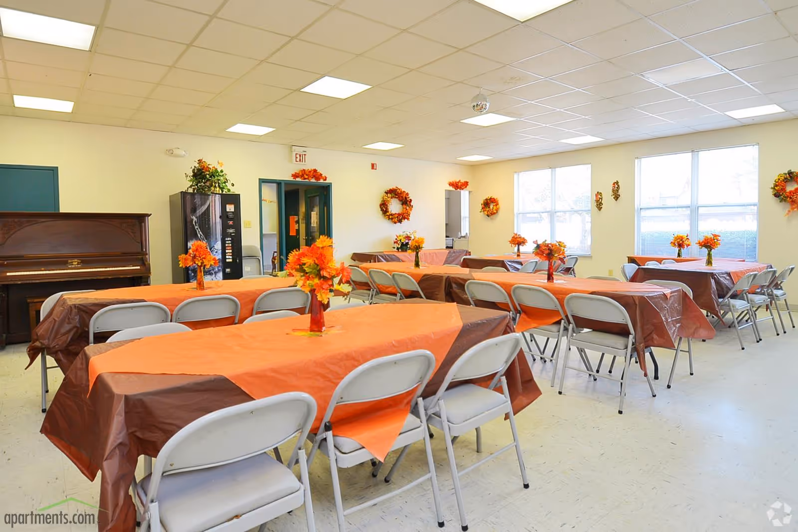 A bright community dining room with several rectangular tables covered in orange and brown tablecloths, each decorated with a vase of orange flowers. White folding chairs are arranged around the tables. The room has large windows letting in natural light, a piano against one wall, a vending machine, and autumn-themed wreaths and decorations on the walls.