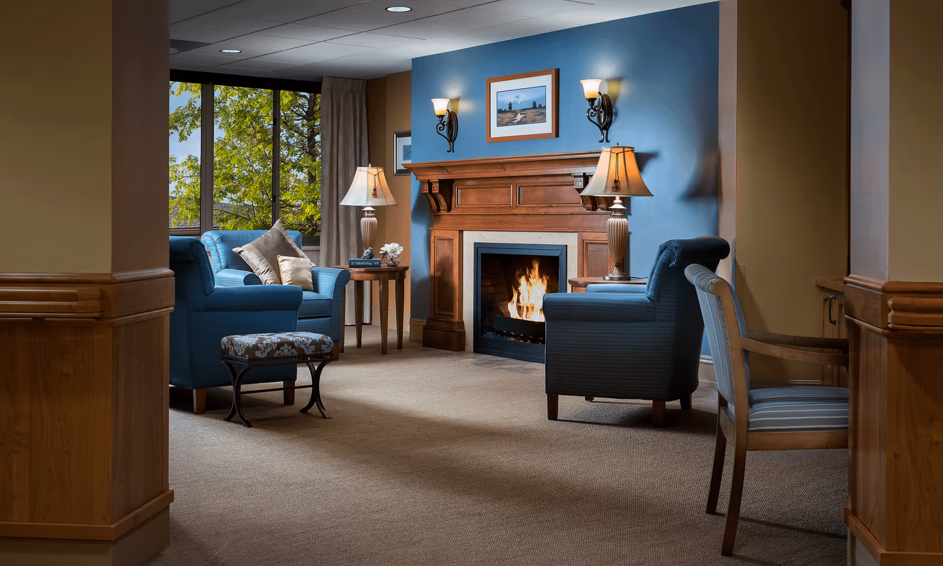 A cozy living room area with a lit fireplace set in a wooden mantel against a blue accent wall. Two blue upholstered armchairs with pillows face the fireplace, accompanied by a small patterned ottoman and a wooden chair with striped upholstery. Two table lamps with beige shades sit on wooden side tables flanking the fireplace. A large window with beige curtains reveals green tree foliage outside.