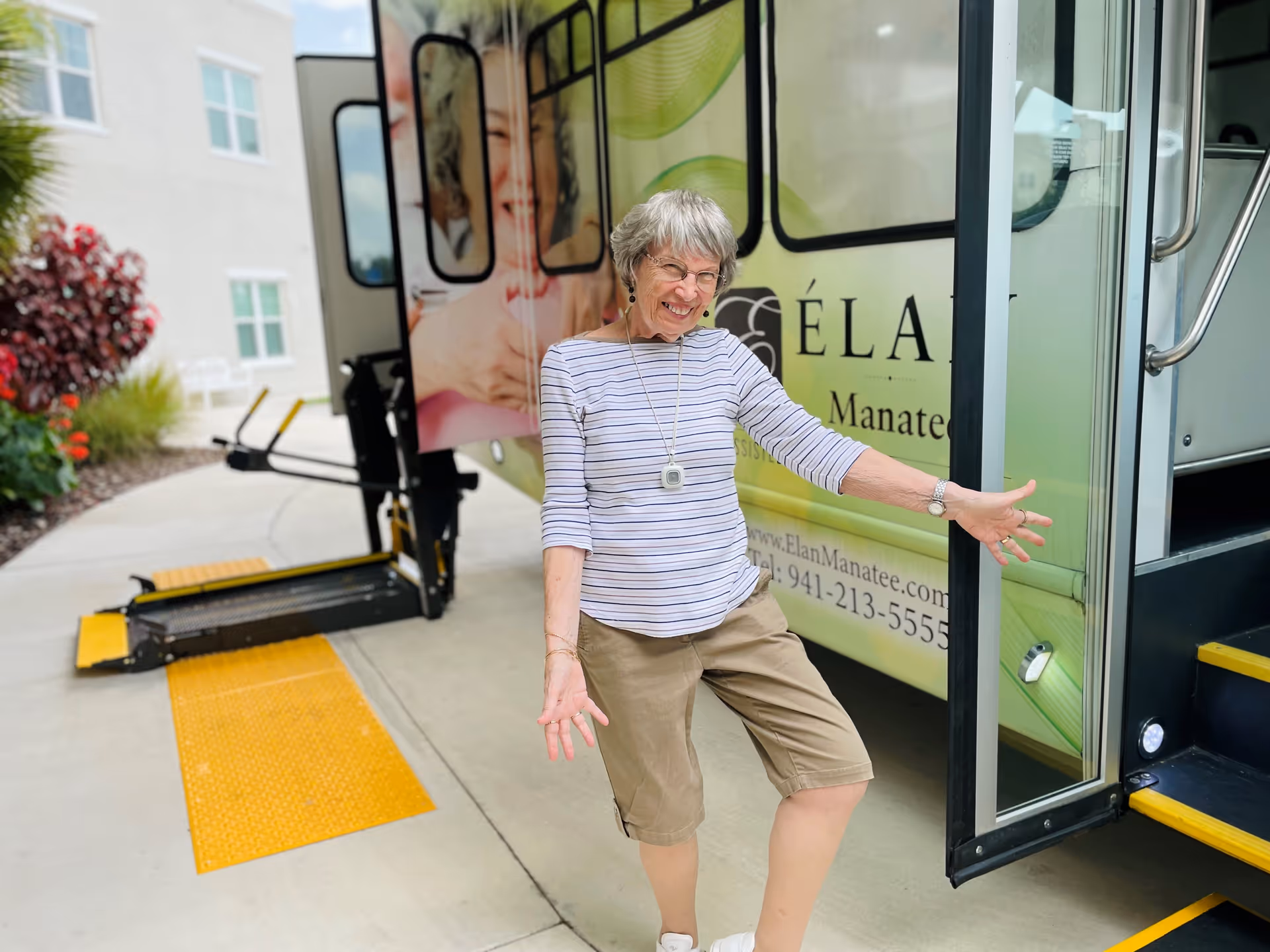 An elderly woman with gray hair and glasses, wearing a striped long-sleeve shirt and khaki shorts, is smiling and posing playfully in front of a vehicle with an open door and a wheelchair lift. The vehicle has signage for Élan Manatee with a website and phone number visible. The scene is outdoors near a building with plants and windows.