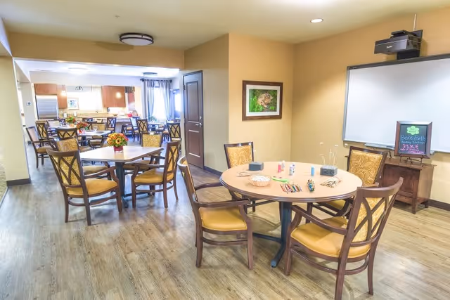 A bright and spacious dining area in Catalina Springs Memory Care with multiple round and square tables surrounded by wooden chairs with yellow cushions. The tables have small flower arrangements and some art supplies. The room has light-colored walls, wood flooring, a framed picture on the wall, and a whiteboard with a small chalkboard sign on a wooden stand.