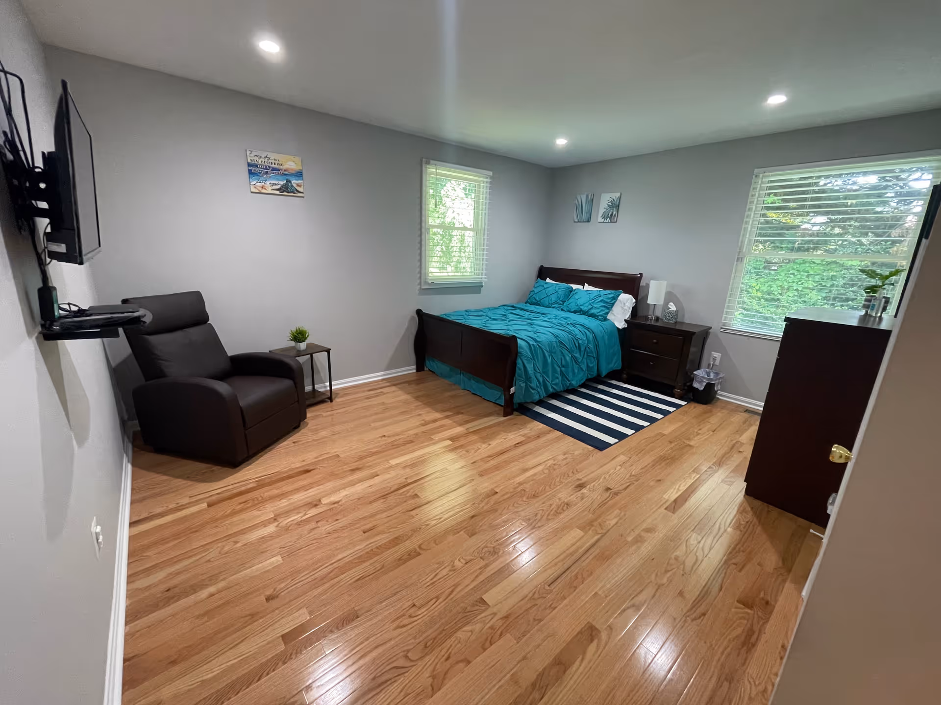 A clean and bright bedroom with light gray walls and wooden flooring. The room features a dark wooden bed with teal bedding, a matching nightstand with a lamp, a striped rug under the bed, a dark recliner chair with a small side table and a potted plant, a wall-mounted TV, two windows with white blinds letting in natural light, and a dark wooden dresser with a small plant on top.