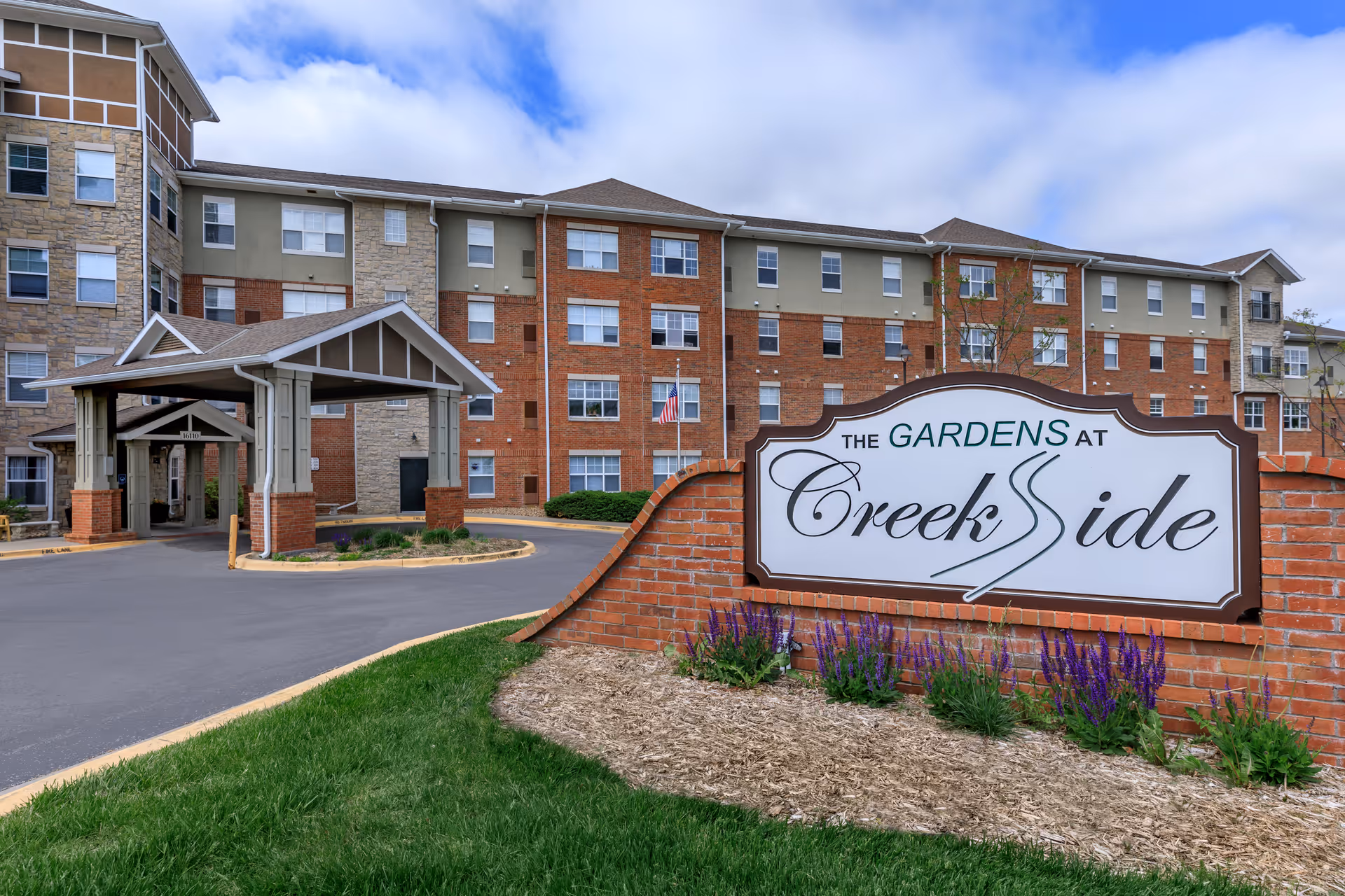 Exterior view of The Gardens at Creekside senior living facility showing a multi-story building with a covered entrance, a brick sign with the facility name, green grass, and purple flowers under a partly cloudy sky.