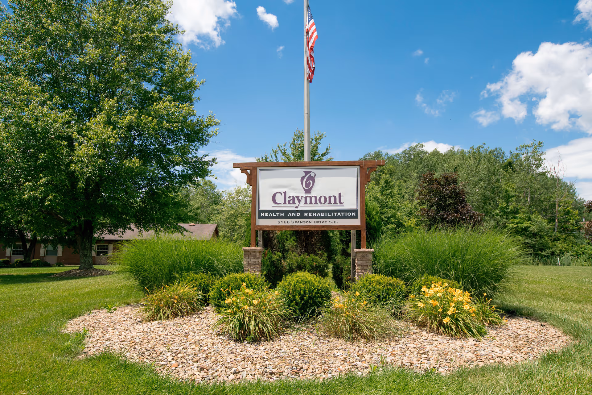 Wooden sign reading 'Claymont Health and Rehabilitation' stands amid landscaping with an American flag, lawn, and trees in the background.