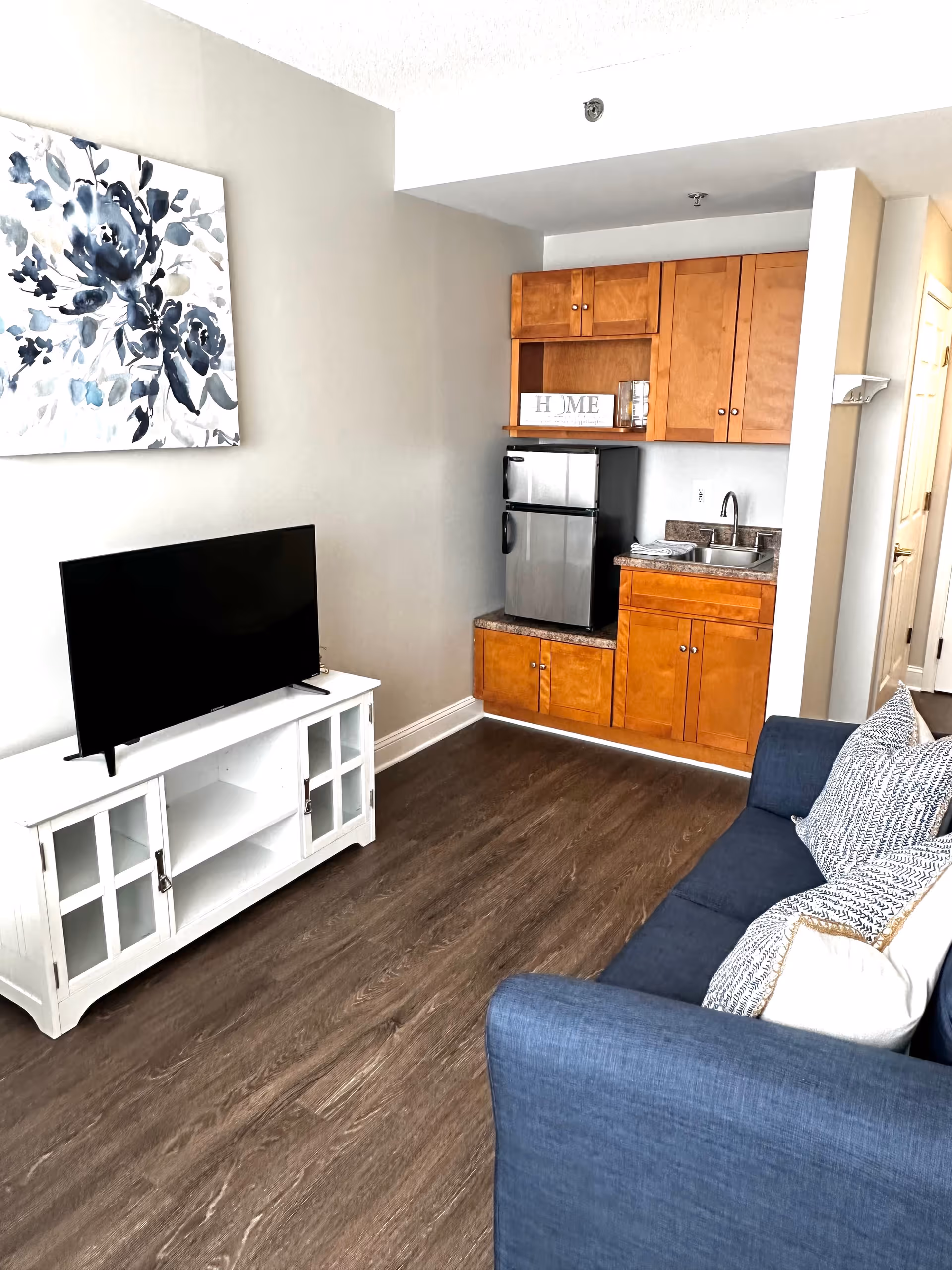 A small living area with a blue couch adorned with patterned pillows, a white TV stand holding a flat-screen television, and a kitchenette featuring wooden cabinets, a small refrigerator, a sink, and a decorative sign that reads 'HOME'. A floral painting hangs on the wall above the TV stand.