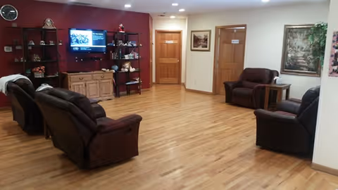 A spacious living room with wooden flooring, featuring several brown recliner chairs arranged around the room. There is a wooden cabinet with shelves holding decorative items and a wall-mounted TV. Two wooden doors and framed artwork are visible on the walls.