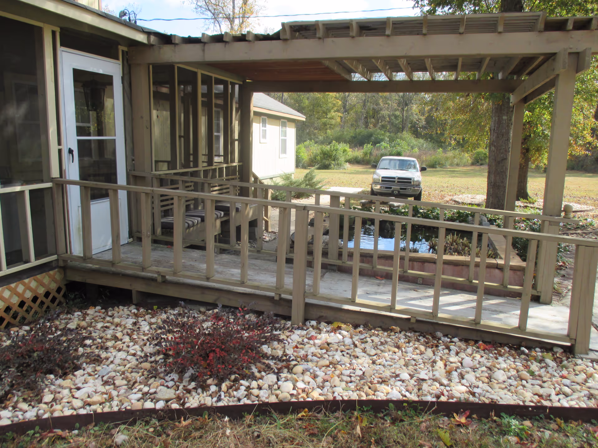 Outdoor wooden patio with a ramp and railing, a small pond with plants, and a white door leading inside. There is a white truck parked on a grassy area in the background with trees and bushes.